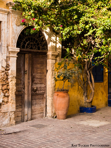 An old, wooden door is flanked by an antique, clay pot and a tree.